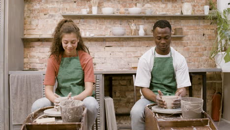 front view of employees wearing green apron modeling ceramic pieces on potter wheel in a workshop while talking to each other 2