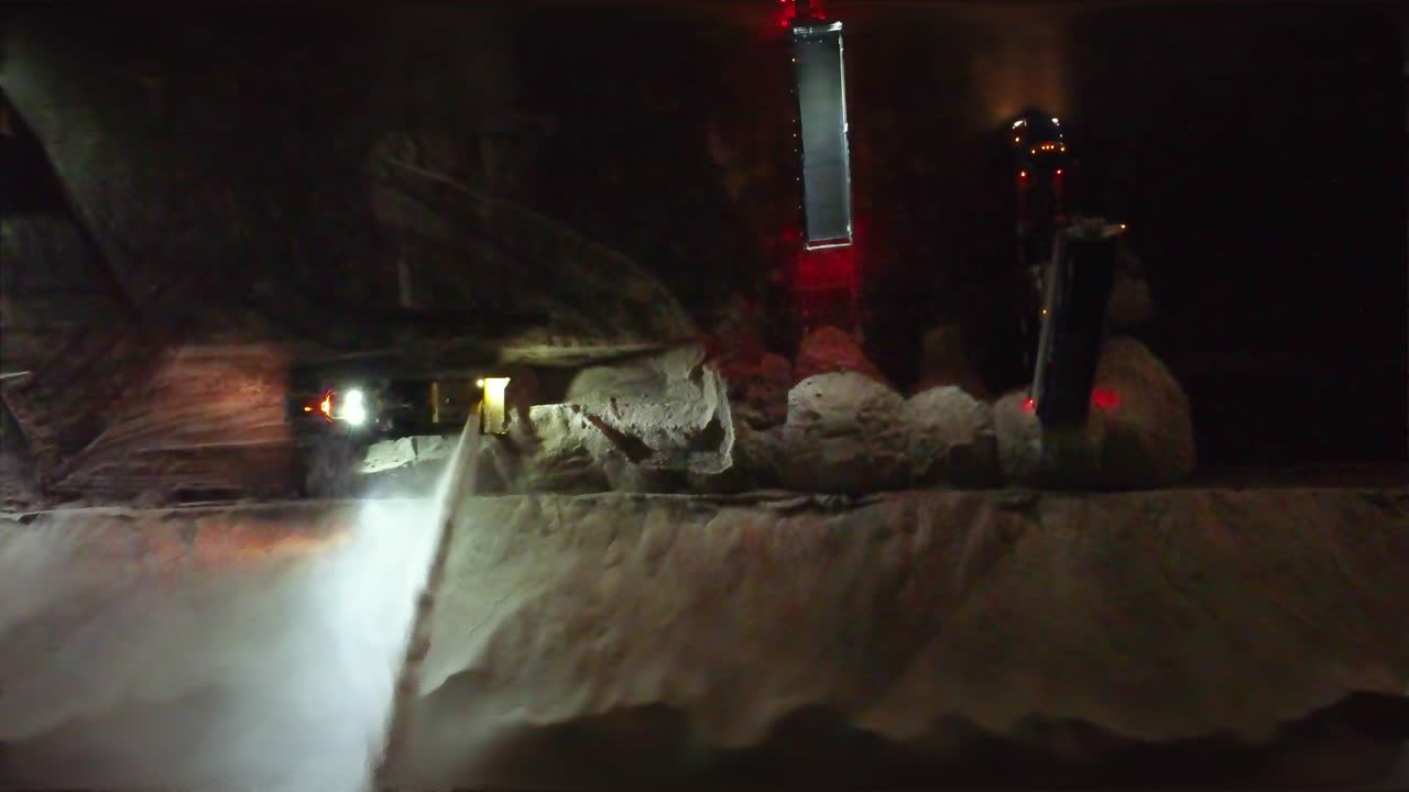 Large commercial snow blower works at night next to semi trucks offloading in empty lot, drone top down