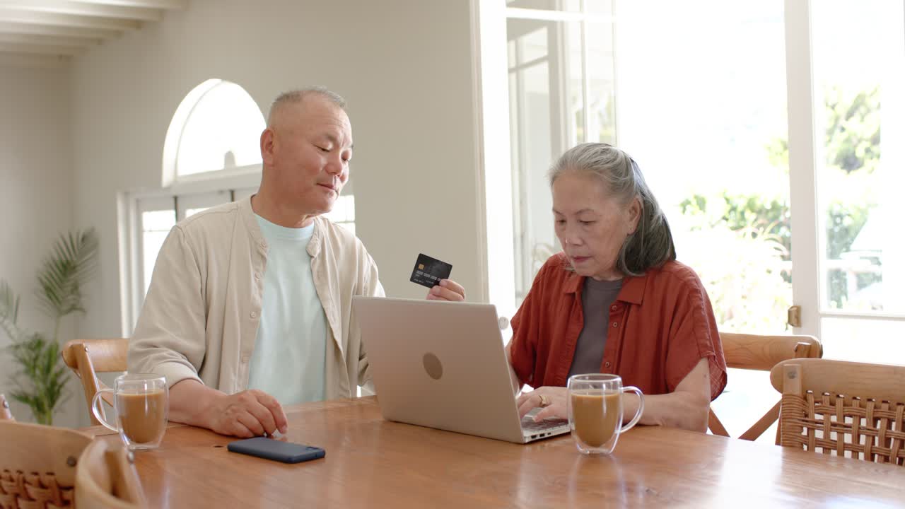 テーブルに座るシニアカップル、男性はクレジットカードを持ち、ラップトップでオンライン決済を行う女性を促している。
