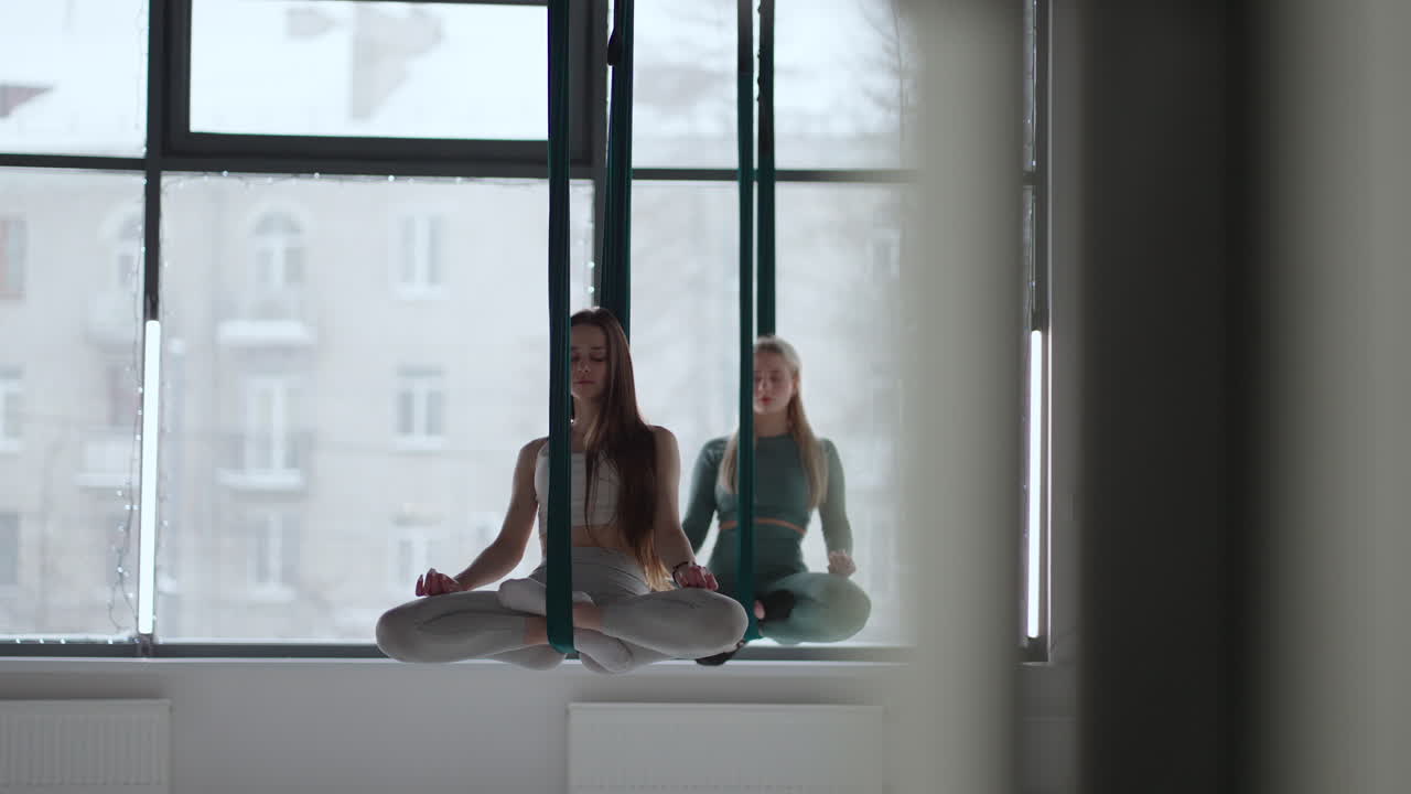 Two young women swing on hammocks and meditate concentrating on their breath. Aero Yoga