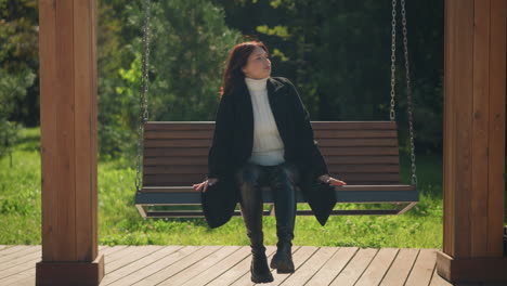 young woman adjusts her hair while seated on wooden swing in sunny park, wearing stylish black coat and boots, surrounded by lush greenery, with serene expression and hand resting on swing