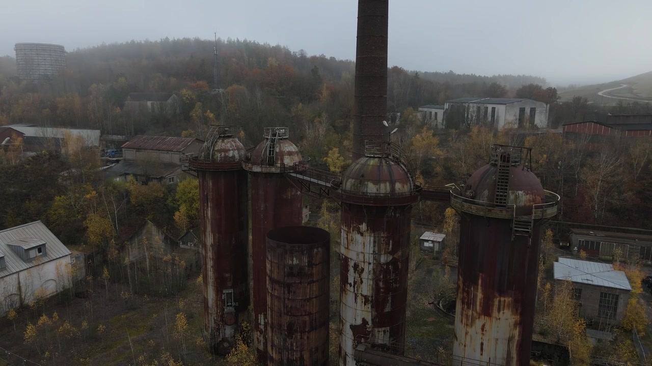 Il drone passa sotto l'altoforno e curva verso il basso oltre serbatoi e silos nella fitta nebbia.