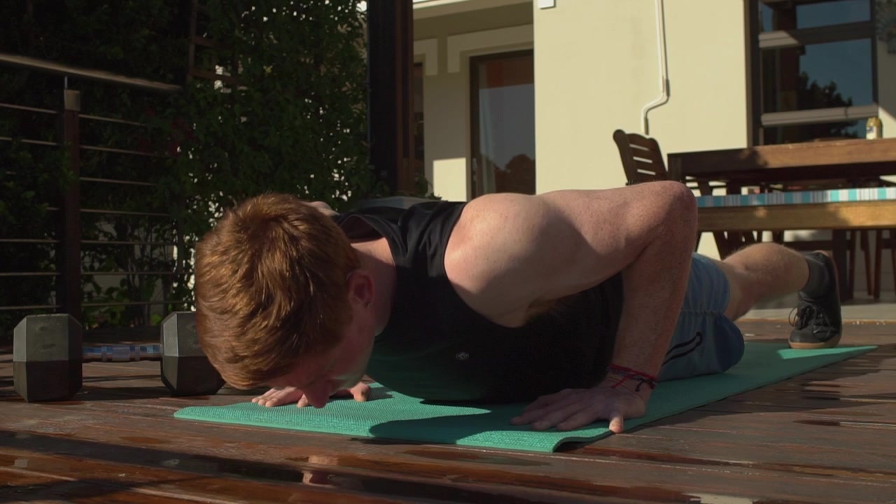 Fit young man with red hair does standard push ups on a green yoga mat over a wooden deck, training his chest and triceps outside in the sun. Static low angle medium view.