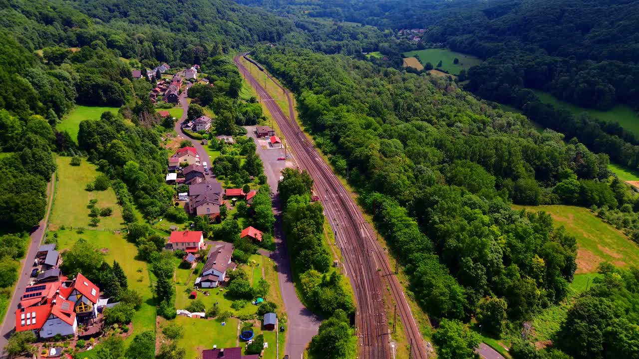 Aerial View of Village with Colorful Rooftops Nestled Beside Multiple Railway Tracks Surrounded by Lush Green Forests