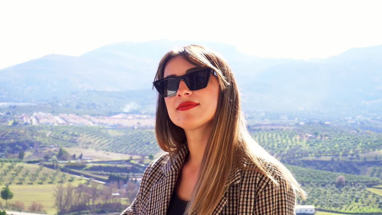 Caucasian girl with sunglasses, straight hair and red lips, touching her hair against the background of some fields and the horizon