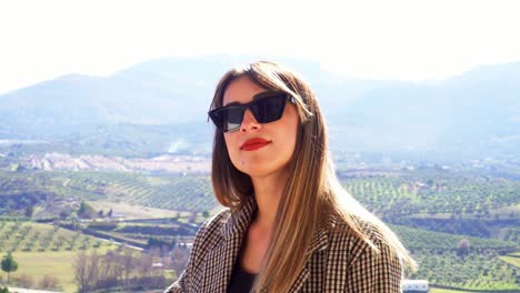 chica caucásica con gafas de sol, pelo lacio y labios rojos, tocándose el pelo contra el fondo de algunos campos y el horizonte