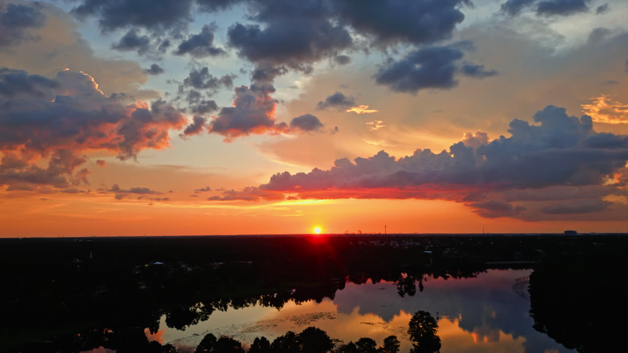Brilliant orange and red sunset colors paint the sky over still water and forested edge, heavenly aerial backdrop background, aerial roll left across the river