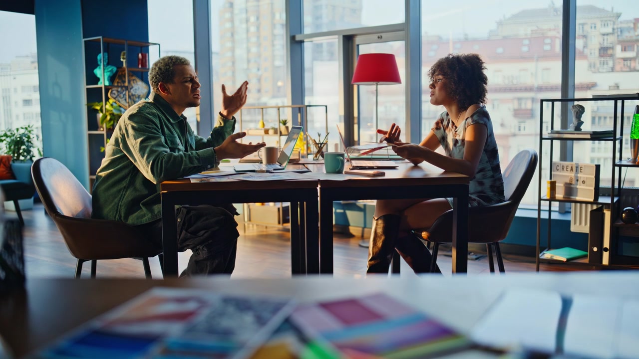 Coworkers arguing across desk. Mixed race pair heated discussion modern office.
