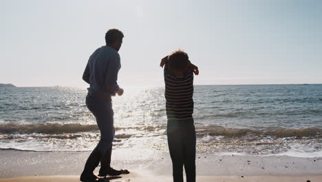 father teaching son to skim stones as they walk along beach by breaking waves together