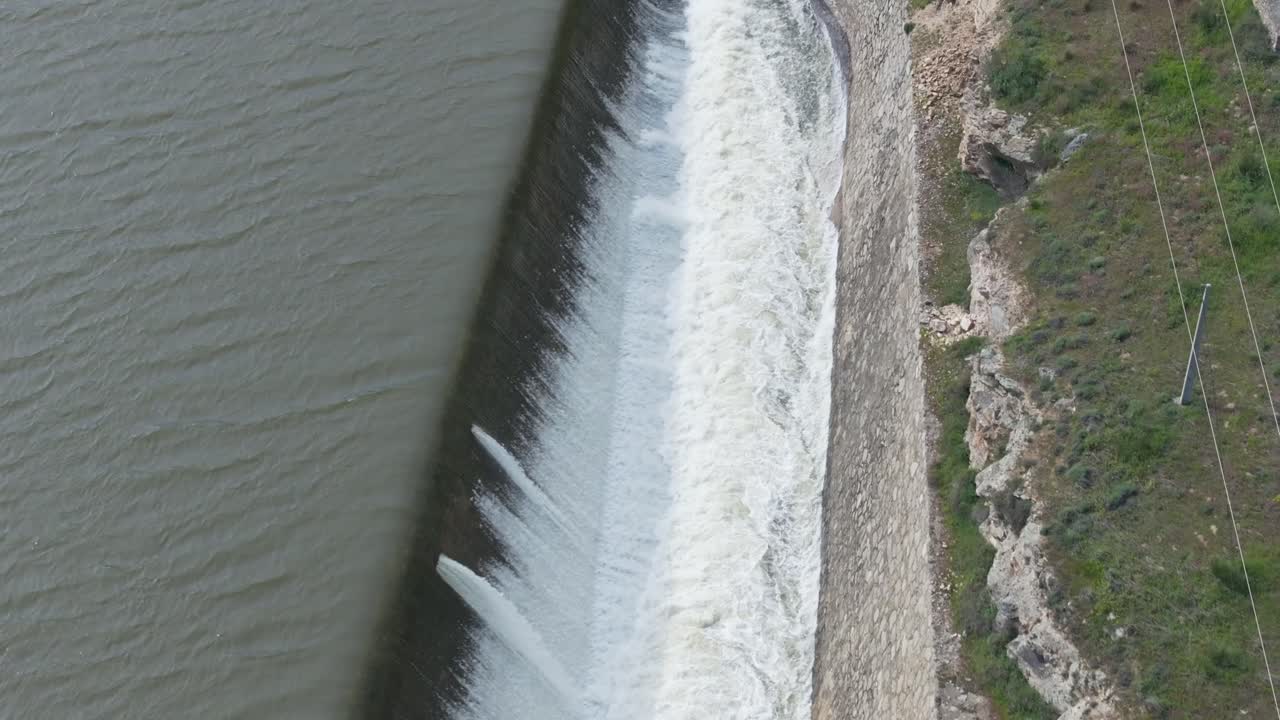 "Reverse drone flight over a dam's lateral spillway, capturing the cascading water, canal, bridge, and powerful river release. Gradually ascending for breathtaking views of nature's force."