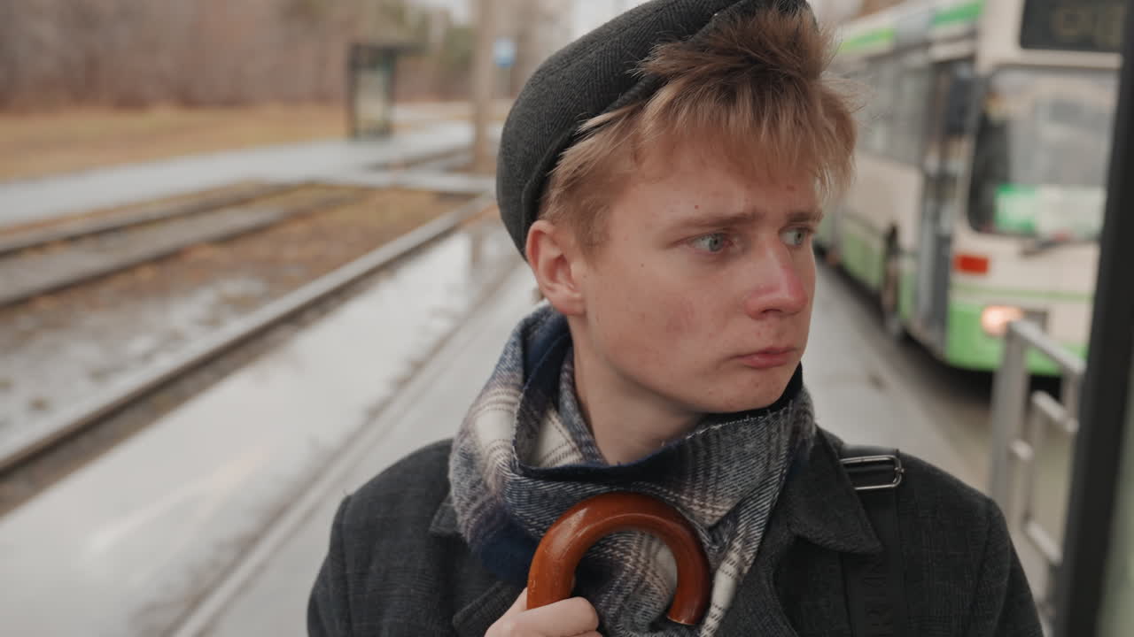 Young white creator poet by tracks watching rain and passing buses, umbrella tucked under arm, notebook closed, reflective expression, slick rails and puddles, distant headlights, cool muted palette