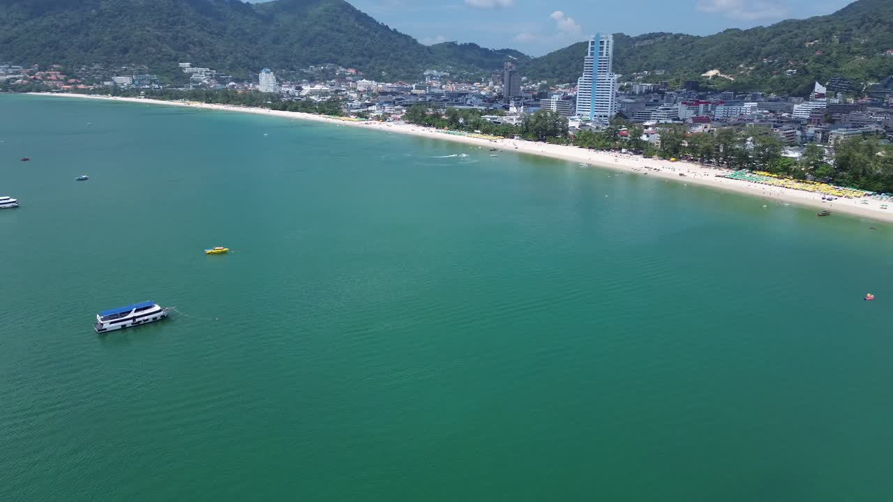 Un drone si alza dietro una roccia, svelando la splendida spiaggia di Patong con acque turchesi e bagnanti che si godono la costa soleggiata di Phuket, Thailandia.
