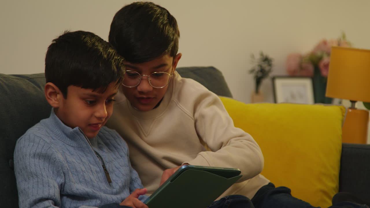 Two Young Boys Sitting On Sofa At Home Playing Games Or Streaming Onto Digital Tablet Together