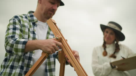 a close-up of a craftsman wearing a plaid shirt and hat as he sets up a wooden tripod in a grassy field. in the background, a woman smiles warmly at him, adding a sense of connection and shared