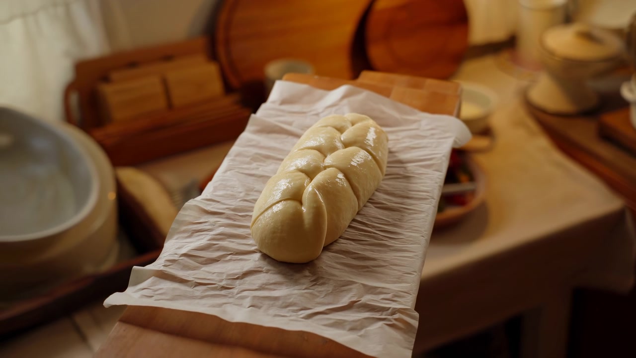 Um pão trançado de massa crua repousa sobre papel manteiga amassado numa bancada de madeira, com utensílios de panificação e tigelas dispostos ao redor sob luz interior quente.