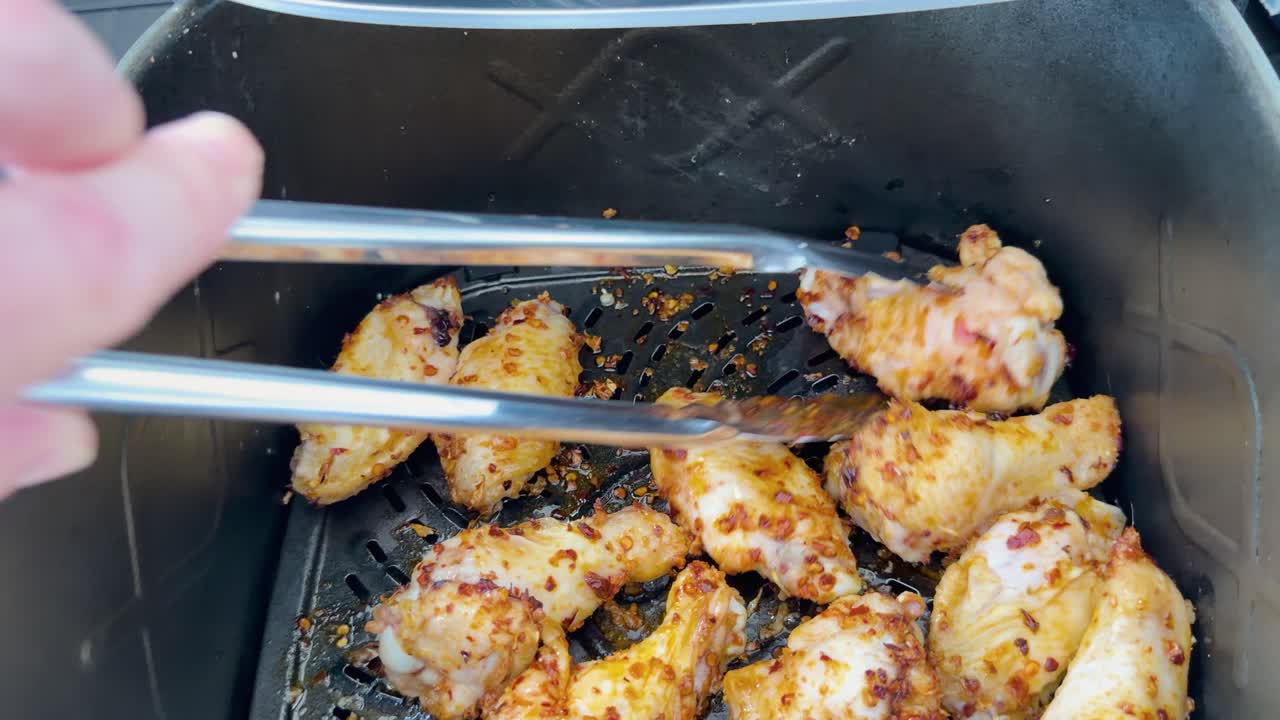 Hand turns spiced chicken wings with metal tongs inside air fryer under bright natural light