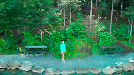 Drone-shot-going-up,-revealing-older-sporty-woman-waving-by-a-beautiful-lake-in-nature