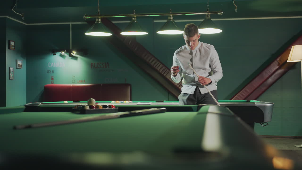색 셔츠와 회색 바지를 입은 플레이어는 초록색 빌리어드 테이블에 앉아 있으며, 집중된 표정으로 큐 스틱을 들고 있습니다. 배경에는 배열된 블리어드 공이 있는 다른 빌리어트 테이블이 보입니다.
