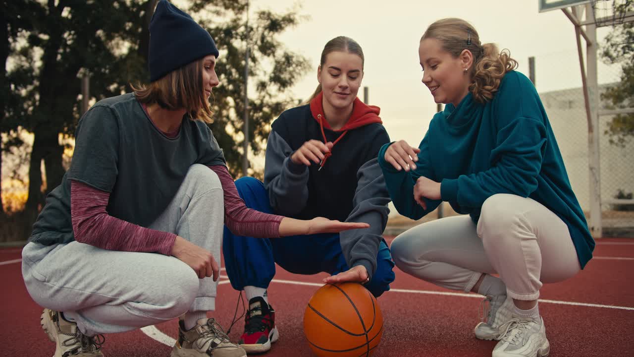 un trío de chicas rubias felices en el mismo equipo de baloncesto pusieron sus manos juntas y las levantaron en un signo de unidad cerca de la pelota de basket en la cancha de basket roja temprano en la mañana