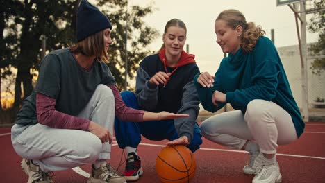 A-trio-of-happy-blonde-girls-on-the-same-basketball-team-put-their-hands-together-and-raise-them-up-in-a-sign-of-unity-near-the-basketball-ball-on-the-red-basketball-court-early-in-the-morning