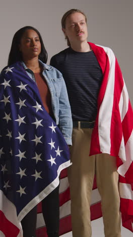 retrato vertical de estudio de video de una pareja multicultural envuelta en la bandera estadounidense celebrando el 4 de julio, día de la independencia