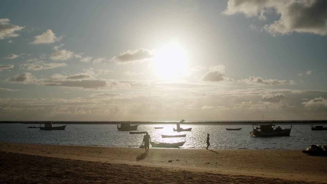 황금시간대에 정박한 배로 가득 찬 작은 만에서 낚시를 위해 작은 노젓는 배를 준비하는 두 남자의 실루엣과 모래사장을 걷는 사람