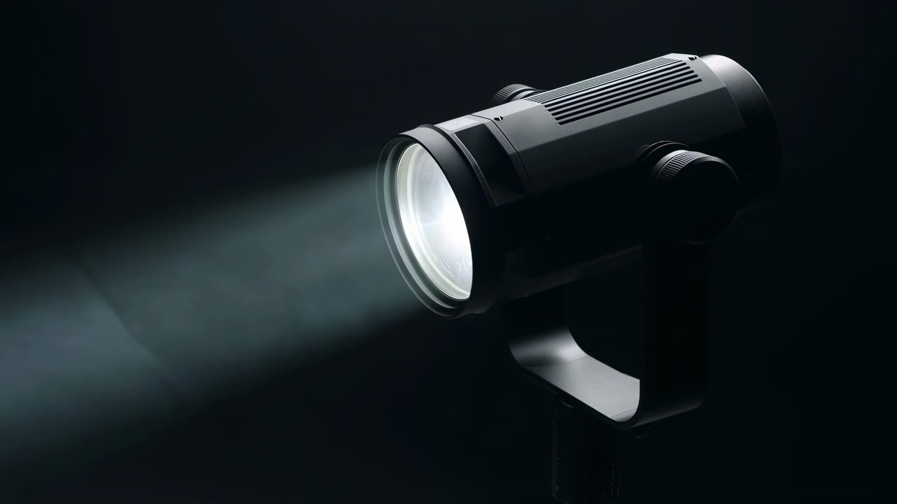 A partially lit cylindrical spotlight fixture stands against a dark background, casting a soft directional beam with visible haze and dramatic contrast from a side view