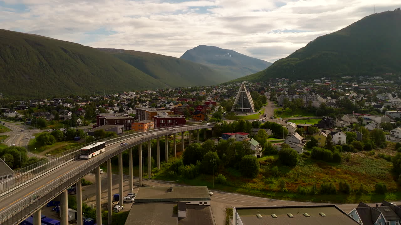 Autobus turistico sul Ponte di Tromsø con l'iconica Cattedrale Artica e le montagne verdi sullo sfondo, Tromsø, Norvegia. Ripresa aerea all'indietro.