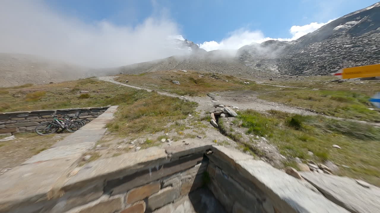 FPV drone follows two mountain bikers descending steep, exposed alpine trails above Saas-Grund and Saas-Fee on a clear bluebird summer day, showcasing epic terrain and speed