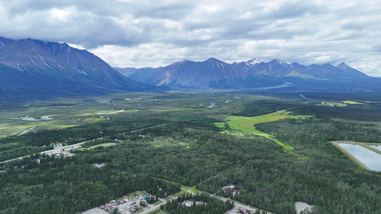 Majestic mountain range overlooks vast green valley with winding river and scattered communities under dramatic cloudy sky in Alaska