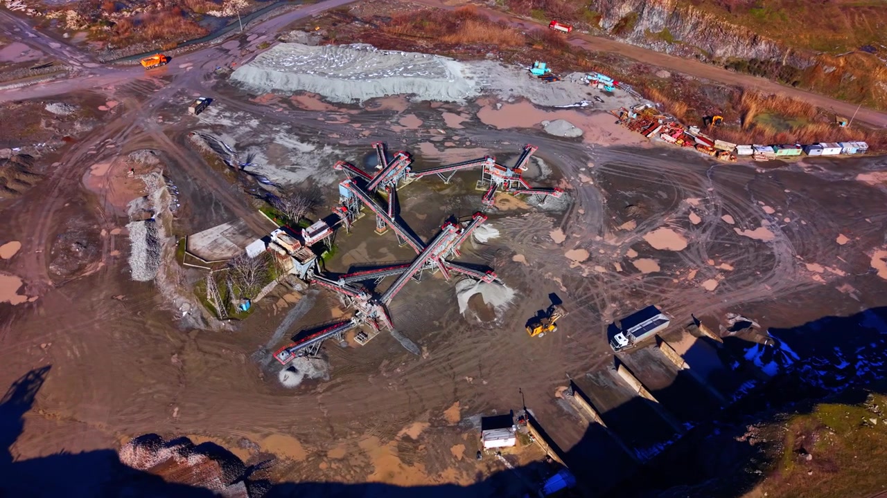 High angle aerial view of a stone crushing plant conveyor system. Overhead look at connected sorting belts, gravel stockpiles, loaders and muddy ground.