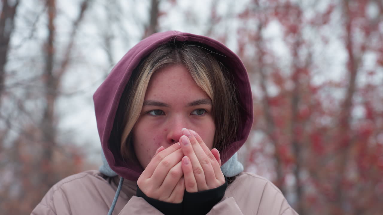 Frau in Winterkleidung steht in der Kälte und bläst warme Luft in die Hände, um sich zu wärmen, Kapuzenjacke und Lagenlook betonen das kalte Wetter, verschwommener Hintergrund aus Bäumen mit roten Beeren.
