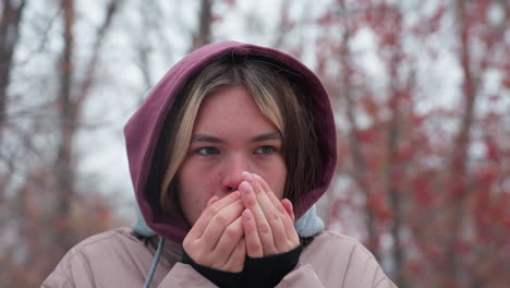 woman in winter outfit standing in cold, blowing hot air into hands for warmth, hooded jacket and layered clothing highlight chilly weather, blurred background of trees with red berries