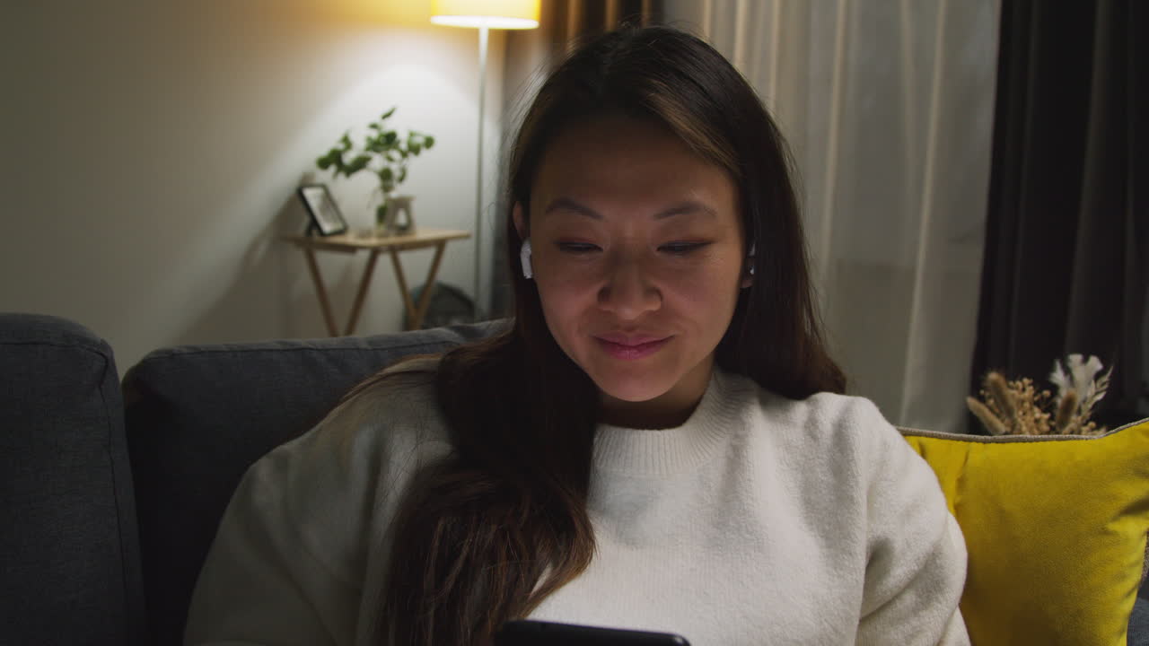 소파에서 집에서 저녁을 보내는 여성, 무선 이어폰을 착용하고 휴대전화에서 음악이나 캐스트를 스트리밍합니다.