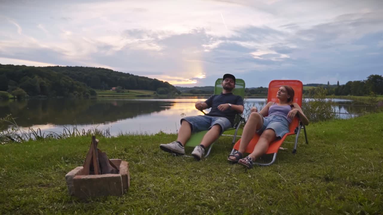 Coppia seduta su sedie da esterno vicino a un lago calmo con un falò al tramonto, che chiacchiera e si gode la tranquilla atmosfera del campeggio nell'ambiente naturale di Savci, Slovenia.