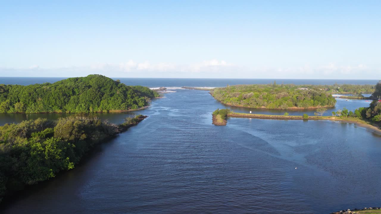 Drone footage captures a serene river flanked by lush greenery under clear skies at Brunswick Heads, NSW, Australia