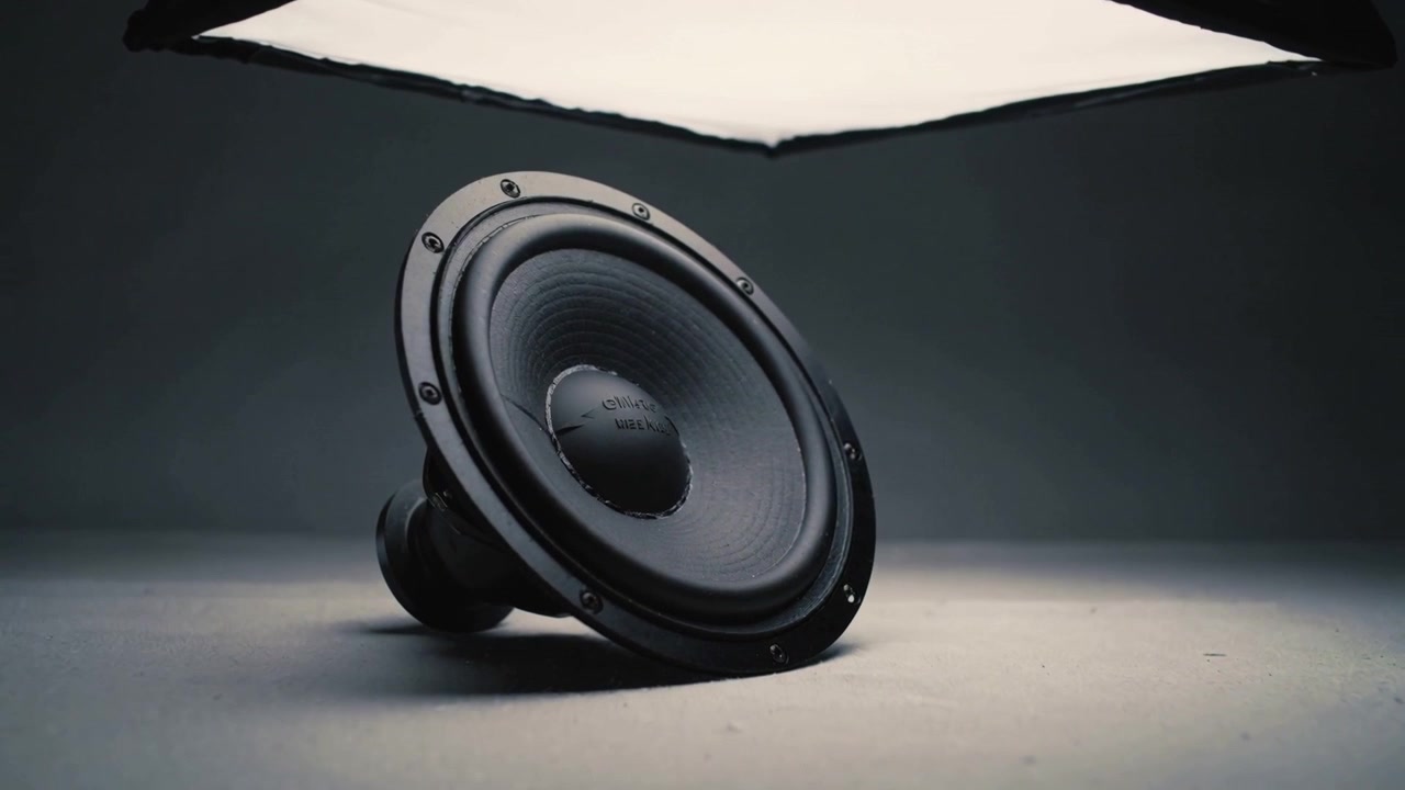 Mostrando un altavoz negro inclinado en el suelo del estudio mientras una caja de luz se desplaza, resaltando la textura del cono.