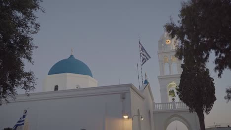 Stolz-Weht-Die-Griechische-Flagge-In-Der-Warmen-Frühen-Nacht-Und-In-Der-Ferne-Sind-Die-Blauen-Kuppeln-Der-Griechisch-orthodoxen-Kirche-Sichtbar,-Sie-Sind-In-Griechenland