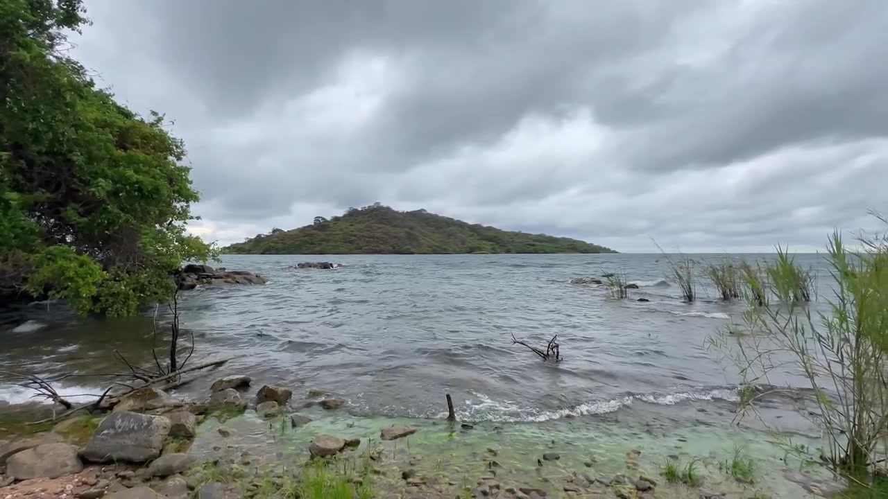 Vlny se jemně vlní o skalnaté pobřeží na jezeře Malawi, obklopené bujnou zelení pod zataženou oblohou.