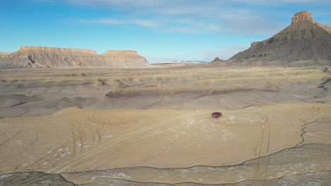 Vista-Aérea-De-La-Camioneta-Roja-Girando-En-Tierra-Seca-Del-Desierto-Con-Formación-Rocosa-De-Fábrica-En-El-Fondo,-Urah,-EE.UU.