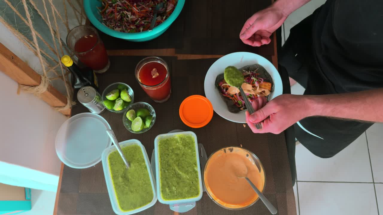 Top down view of man garnishing Baja fish tacos with salsa, surrounded by limes, sauces, and Micheladas