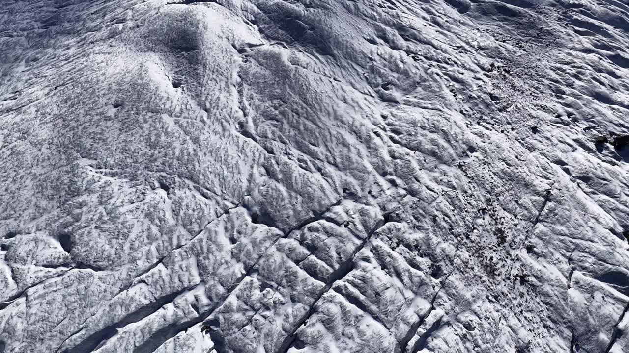 A top-down aerial view of a frozen glacier surface. The crevassed ice texture dominates the foreground, with a snow-capped mountain top or ridge visible in the background. Swiss Alps, Graubünden