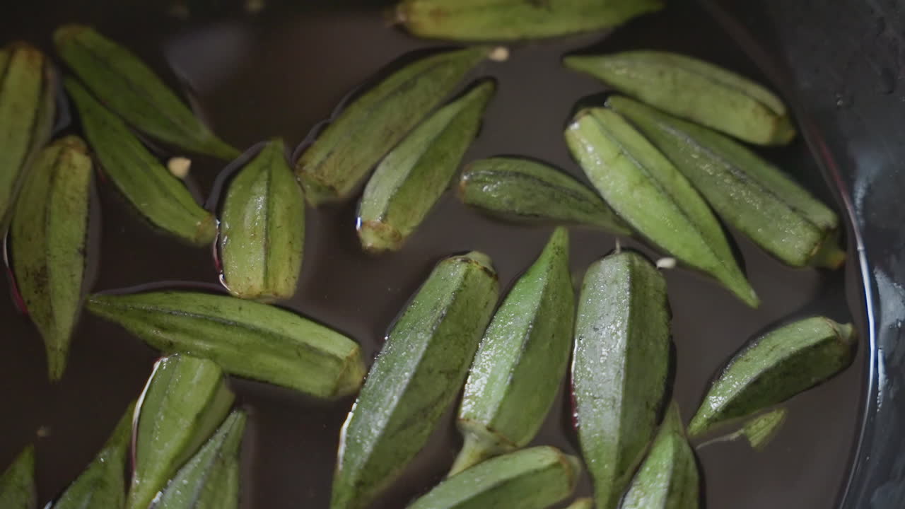 Tampilan dekat buah okra di wajan berkilauan dengan kristal garam dan tetesan air, koki rumahan menyiapkan bahan untuk resep, fokus pada alur sayuran, tekstur, dan komposisi pedesaan, uap tipis dan aroma tanah.