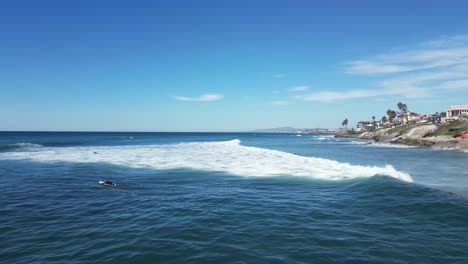 Drone-shot-pulling-backwards-over-sunny-ocean-with-beautiful-blue-and-green-waves-and-surfers-paddling-around