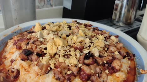 close-up shot of a deep plate with octopus salad in a kitchen, while an elderly woman adds ingredients