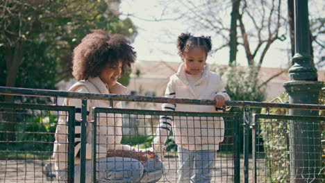 little girl throwing crumbs to wild birds leaning on park fence with mother.