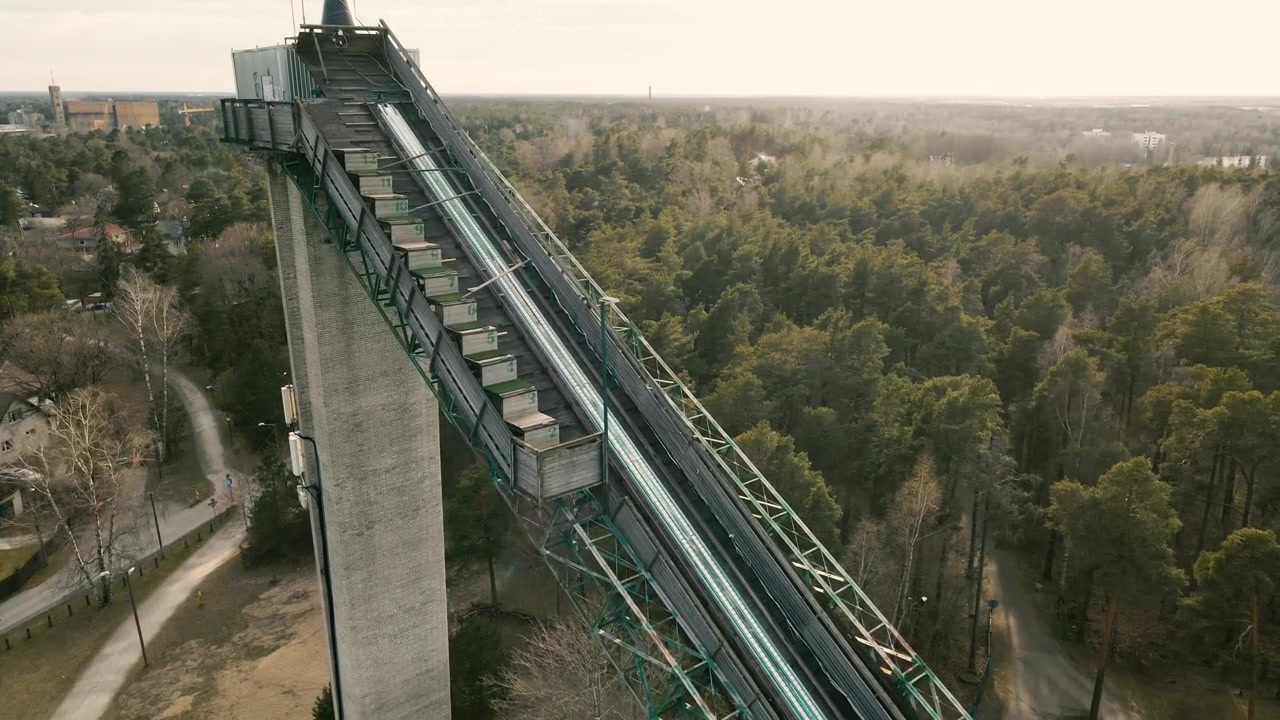 Riprese aeree con drone che affiancano una torre di lancio di un trampolino da sci abbandonato, una vecchia struttura in cemento e acciaio che si erge su una vasta e fitta foresta di pini sotto un cielo coperto.