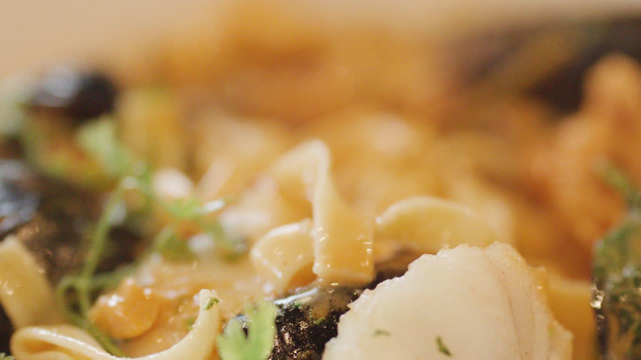 Macro view of hot seafood chowder with mussels, scallops, noodles, and fresh herbs, as a hand stirs with a spoon under warm lighting