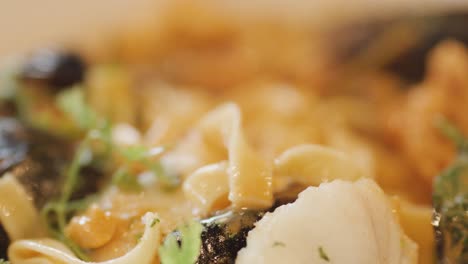 macro view of hot seafood chowder with mussels, scallops, noodles, and fresh herbs, as a hand stirs with a spoon under warm lighting