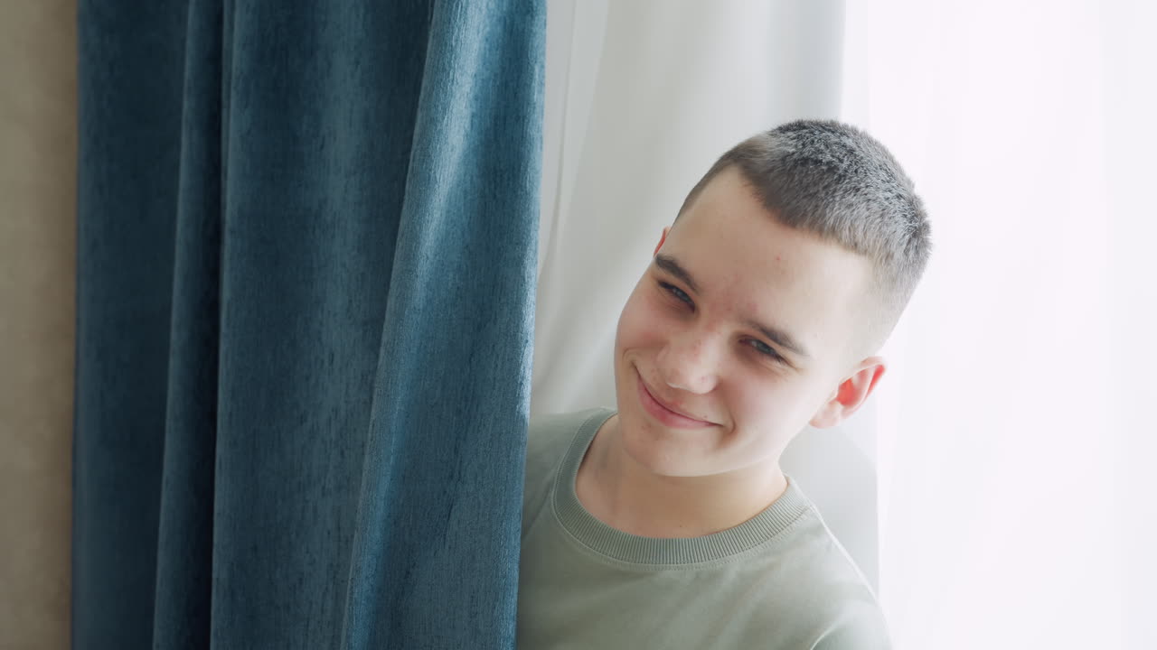 Boy partially concealed behind thick blue curtain with white clothing in brightly lit room as sunlight streams through sheer white drapes, capturing playful secretive moment of hiding
